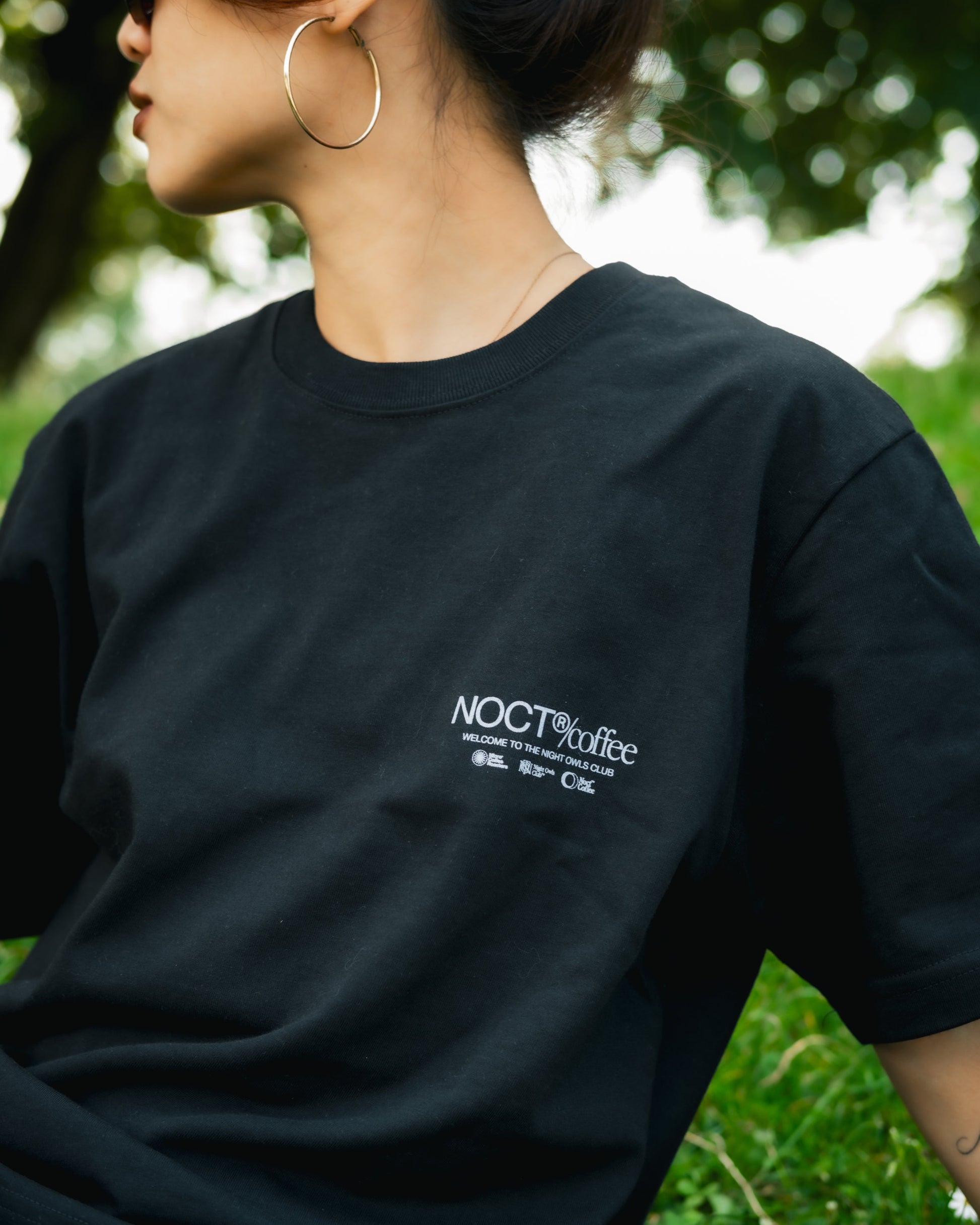 A person outdoors wearing a black oversized T-shirt with a small white 'NOCT coffee' logo on the left chest, paired with light gray sweatpants. The background shows blurred green grass and foliage.