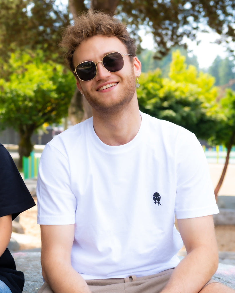 A man wearing a white crewneck t-shirt with a small black "nyx" noct owl logo on the left chest, sitting outdoors in a park-like setting with green trees in the background.