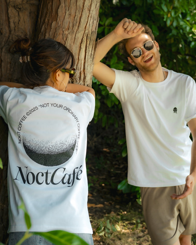 Two models wearing white NoctCafé t-shirts in an outdoor forest setting. The model on the left shows the back of the white t-shirt featuring a large black coffee bean graphic and the text 'NoctCafé'.